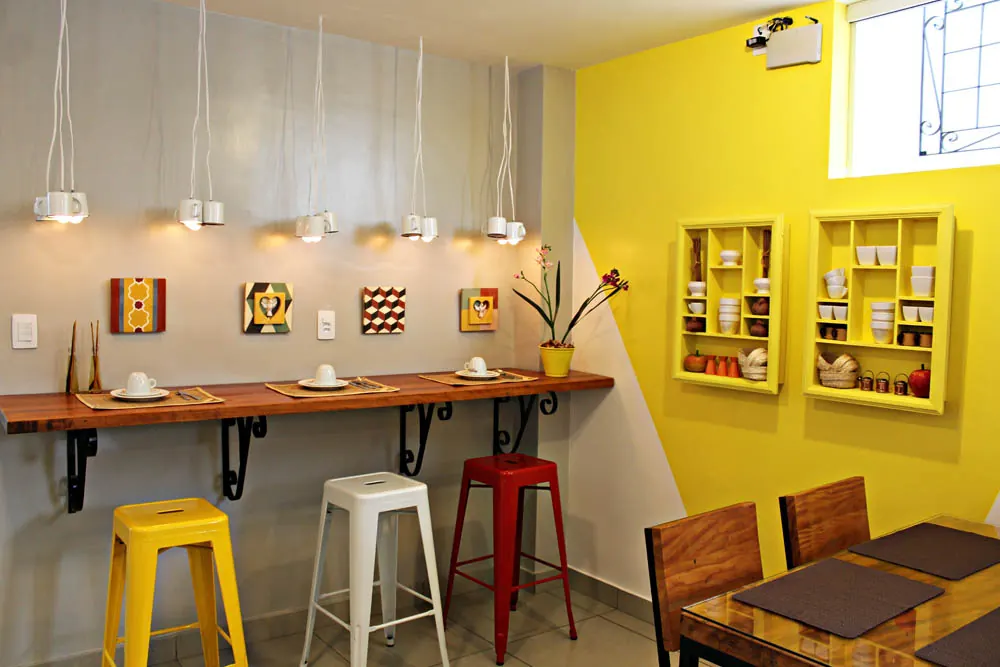 Sala do café da manhã com parede amarela, bancos coloridos e quadros de artesanato